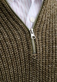 Strickpullover aus olivgrüner Wolle mit sichtbarer Rippenstruktur, versehen mit einem silbernen Reißverschluss und einem darunterliegenden weiß gestreiften Hemdkragen.