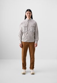 Light gray button-up shirt with two chest pockets, paired with brown rolled cuff trousers and beige sneakers on a plain background.