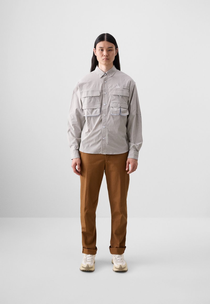 Light gray button-up shirt with two chest pockets, paired with brown rolled cuff trousers and beige sneakers on a plain background.