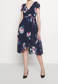 Floraalinen kietaisumekko tummansinisestä kankaasta, jossa on lyhyet puffihihat, vyötäröllä solmittava nauha, epäsymmetrinen helma ja moniväriset kukkakuviot.