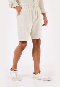 Pantaloni corti beige chiaro in tessuto testurizzato con vita elasticizzata e cordoncino; abbinati a sneakers bianche con dettagli beige.