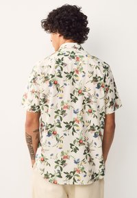 Camicia a maniche corte in tessuto leggero con sfondo crema e motivi floreali e fruttati colorati. Colletto con bottoni, vestibilità comoda.