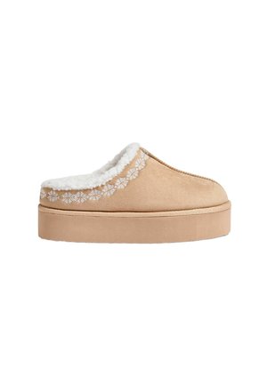 Beige ruskind slip-on hjemmesko med tyk sål, hvid fleeceforing og dekorative hvide syninger nær ankelkanten.