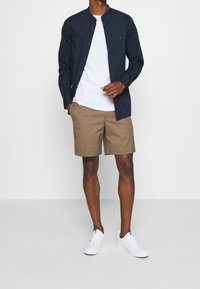 Camicia blu navy con bottoni sopra una t-shirt bianca, abbinata a pantaloncini beige e scarpe da ginnastica bianche. Tessuto liscio, design casual e vestibilità moderna.