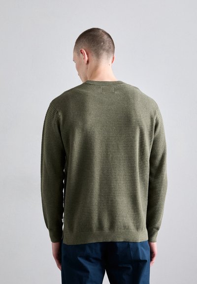 Pull en laine vert olive avec un motif texturé, un col et des poignets côtelés, et un ourlet droit. Porté avec un pantalon bleu marine.