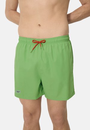 Hombre vistiendo pantalones cortos de baño verde brillante con un cordón rojo, de pie contra un fondo blanco liso.