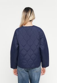 Person mit langen blonden Haaren, der eine gesteppte marineblaue Jacke und blue Jeans trägt, von hinten gegen einen schlichten weißen Hintergrund blickend.