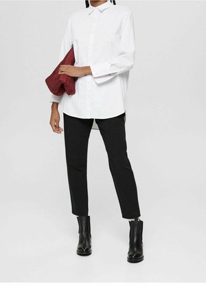 Osoba ubrana w białą koszulę zapinaną na guziki, czarne spodnie, czarne botki za kostkę, trzymająca złożoną bordową kurtkę pod pachą.