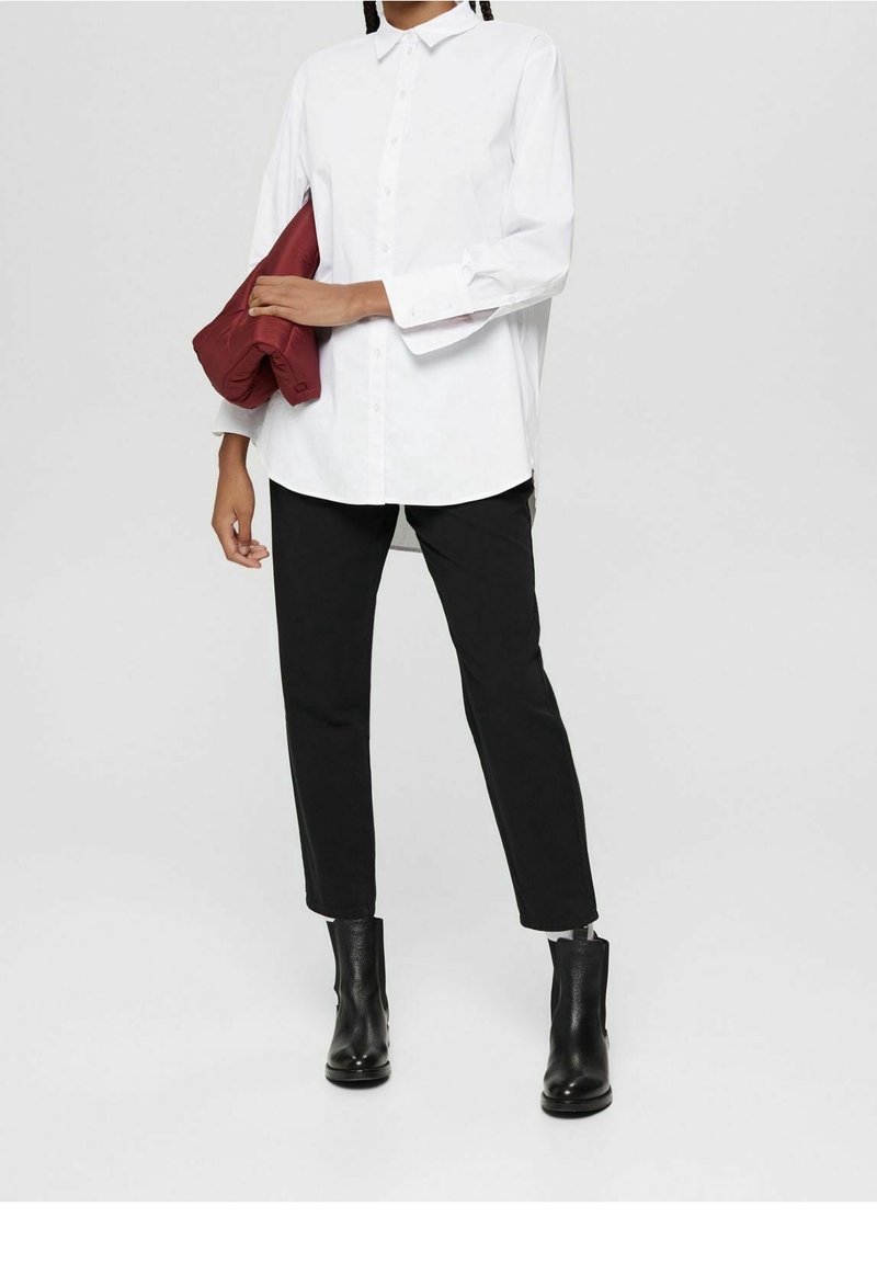 Personne portant une chemise blanche boutonnée, un pantalon noir, des bottines noires, tenant une veste bordeaux pliée sous le bras.