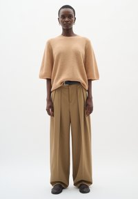 Beige gerippter Strickpullover in lockerer Passform, kombiniert mit hellbraunen, weit geschnittenen Hosen mit Falten. Ein schwarzer Gürtel und dunkle Schuhe runden den Look ab.