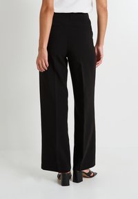 Pantalones de pierna ancha negros con cintura alta, con bolsillos traseros y una textura de tejido suave. Combinados con sandalias de tacón abierto.