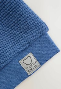 Blue knitted fabric with textured and smooth sections, featuring a small white label with a teddy bear outline and text "LITTLE ONE-365".