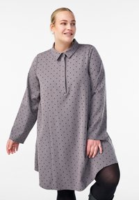 Nainen, joka on pukeutunut harmaaseen pitkähihaiseen mekkoon, jossa on mustia pilkkuja, mustiin sukkahousuihin ja puolipitkät saappaat, seisoo valkoista taustaa vasten.