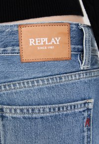 Slitte lys blå denimjeans med et brunt lærmerke som leser "REPLAY SINCE 1981" og røde sømmer på baklommen.