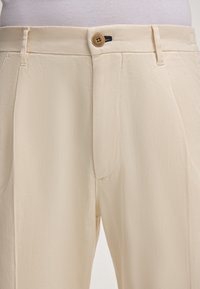 Beige broek met een gladde textuur, voorzien van een knoopsluiting, riemlussen en zijzakken. Op maat gemaakt ontwerp met nette stiksels.