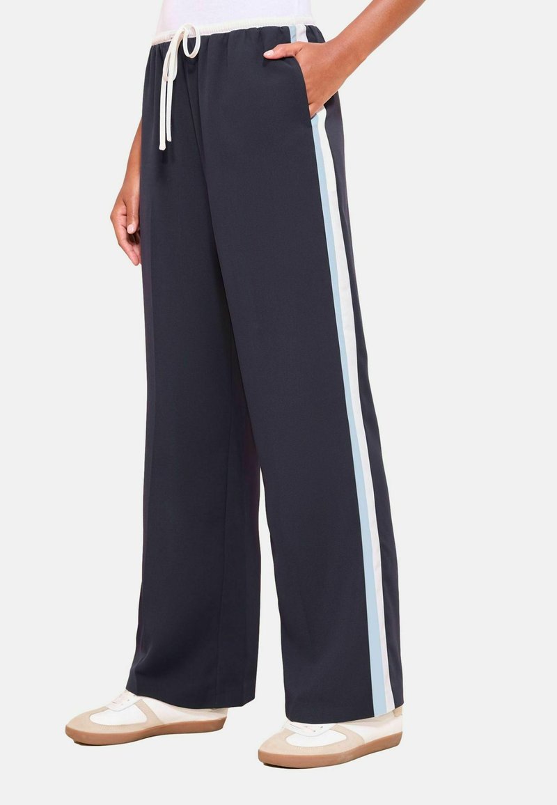 Persoon in donkere wijde broek met witte en lichtblauwe zijkanten en witte sneakers met beige accenten.