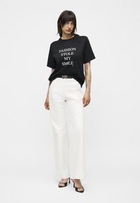 Žena s tamnom kosom nosi crnu majicu s natpisom "FASHION STOLE MY SMILE", bijele široke hlače, crni remen i crne otvorene pete, stoji.