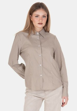 Giovane donna con capelli biondi lisci indossa una camicia beige e bianca a righe con bottoni e pantaloni beige chiaro, posando con una mano dietro la schiena.