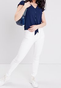 Topp marineblå med knapper, V-hals og korte ermer, kombinert med hvite smale jeans og hvite joggesko. Kuttet design med en knytedetalj.