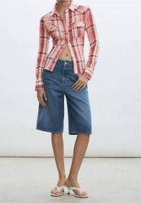 Chemise à carreaux rouges à boutons avec des manches longues, associée à un short en denim bleu Bermuda. Des sandales en plastique blanches complètent la tenue.