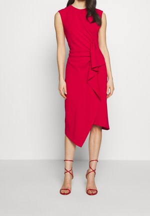 Kobieta ubrana w czerwoną sukienkę midi bez rękawów z drapowanym detalem i asymetrycznym dołem, połączoną z czerwonymi sandałkami na wysokim obcasie z paskami.