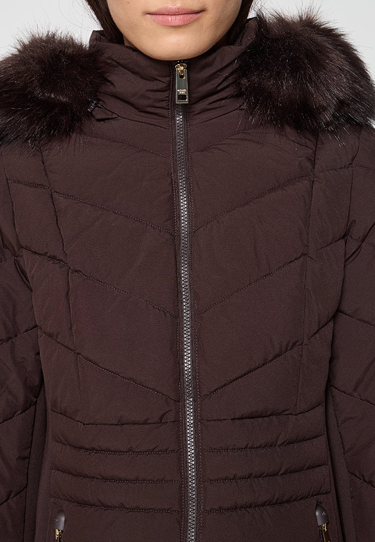 Vrouw in een donkere bruine gevoerde winterjas met rits, voorzien van een kraag met bont en een hoge hals.