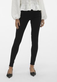 Jeans negros ajustados con una textura suave y un ligero brillo, combinados con tacones negros afilados. La modelo lleva una blusa blanca texturizada.