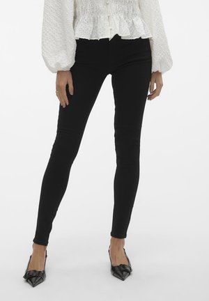 Jeans noirs ajustés avec une texture lisse et un léger éclat, accompagnés de talons aiguilles noirs. Le mannequin porte un haut blanc texturé.