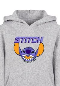 Šedá mikina s farebnou grafikou rozprávkového charakteru s fialovým kožuchom a oranžovými akcentmi. Text znie "STITCH EXPERIMENT 626."
