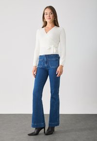 Bluză albă cu rigle, cu un nod la talie, asociată cu blugi evazați bleumarin și pantofi cu vârf ascuțit negri. Texturi fine și design ajustat.