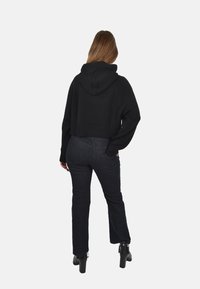 Felpa corta nera con cappuccio regolabile e orlo a costine, abbinata a jeans scuri e stivaletti, vista da dietro.