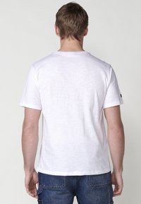 White short-sleeve t-shirt with a round neckline, made of textured fabric. Features a small brown accent at the left shoulder.
