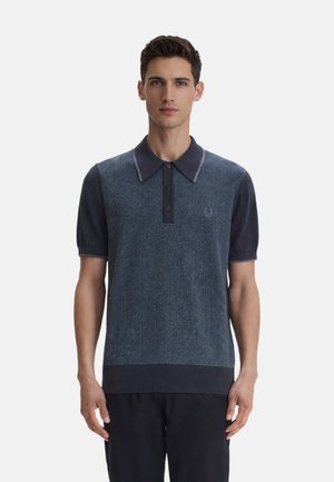 Joven con una camisa tipo polo de manga corta azul con estampado, cuello y tres botones, de pie frente a un fondo blanco liso.