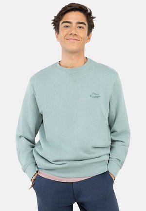 Joven con cabello corto y oscuro, lleva una sudadera azul claro y pantalones azul marino, sonriendo con las manos en los bolsillos sobre un fondo liso.