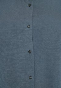 Košulja s dugim rukavima u laganom plavom materijalu s suptilnom teksturom; sadrži šest tamnih dugmadi duž prednjeg dijela.