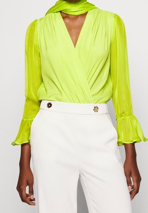 Vrouw draagt een limegroene wikkelblouse met lange mouwen en ruches bij de manchetten en een witte broek met hoge taille en gouden knoopdetails.