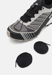 Scarpa sportiva nera e grigia con materiali testurizzati, accenti riflettenti e un sistema di allacciatura integrato. Due lacci rotondi neri affianco a essa.
