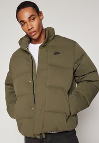 Giacca puffer verde oliva con chiusura a zip, bottoni a pressione e logo nero. Presenta un cappuccio, un design trapuntato e polsini elasticizzati.