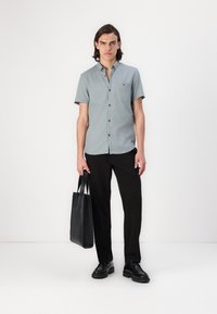 Camisa gris de manga corta con botones y un bolsillo, combinada con pantalones negros y zapatos negros. En la mano izquierda se sostiene un bolso negro.