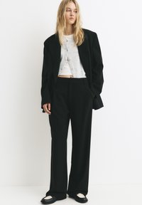 Černý oversized blejzr v kombinaci s kalhotami s vysokým pasem a širokými nohavicemi. Pod ním světle šedý zkrácený cardigan přes bílý top. Hladké textury.