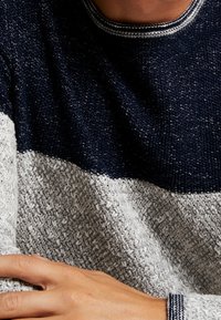 Gruby sweter o grubym splotem, prezentujący poziome paski w kolorze granatowym i jasnoszarym. Okrągły dekolt z subtelnymi akcentami w prążki. Teksturowana tkanina.