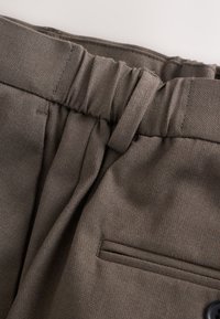 Gros plan sur un pantalon en tissu marron avec une ceinture élastique, une boucle de ceinture et un détail de poche latérale horizontale.