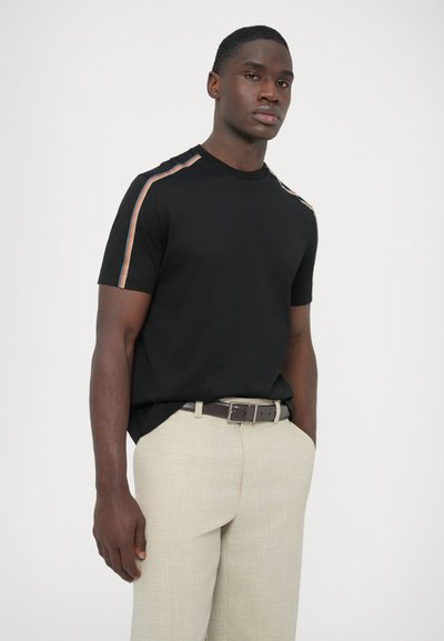 Čierne tričko s krátkym rukávom s viacfarebnými pásikmi pozdĺž ramena, v kombinácii so svetlobéžovými nohavicami a hnedým opaskom.