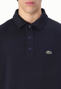Polo de color azul marino hecho de tela de punto, con un cuello clásico, tres botones y un logo bordado verde en el lado izquierdo del pecho.