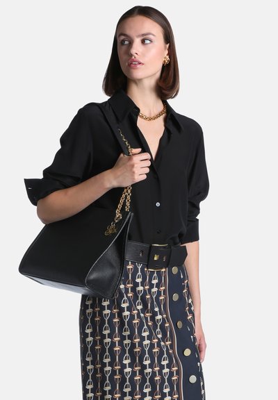Femme portant une chemise noire et une jupe à motifs avec une grande ceinture noire, tenant un sac à main noir avec une bandoulière chaîne dorée, regardant sur le côté.