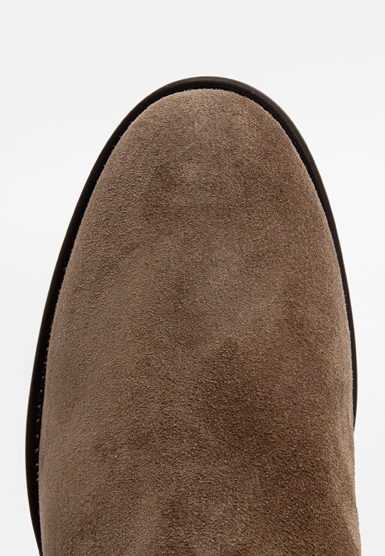 Bottine en suède marron avec un bout arrondi et une fine semelle noire, présentant une texture lisse et sans quincaillerie ni accents visibles.