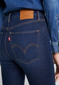 Jeans de mezclilla azul oscuro de cintura alta, con bolsillos traseros prominentes y costuras amarillas. Etiqueta de cuero en la cinturilla que muestra la marca.