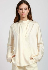 Blusa in seta avorio con colletto alto, maniche lunghe e tasche laterali. Presenta una leggera lucentezza e un accento con cordoncino sul collo.