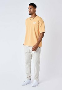 Maglietta a maniche corte color pesco con un motivo testurizzato e orlo a contrasto, abbinata a pantaloni beige chiaro e sneakers bianche.