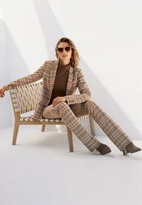 Gecheckte blazer en broek in beige, bruin en crème tinten met een textuurafwerking; gestyled met een ribbelige bruine top en neutrale hakken. Houten lattenbank.